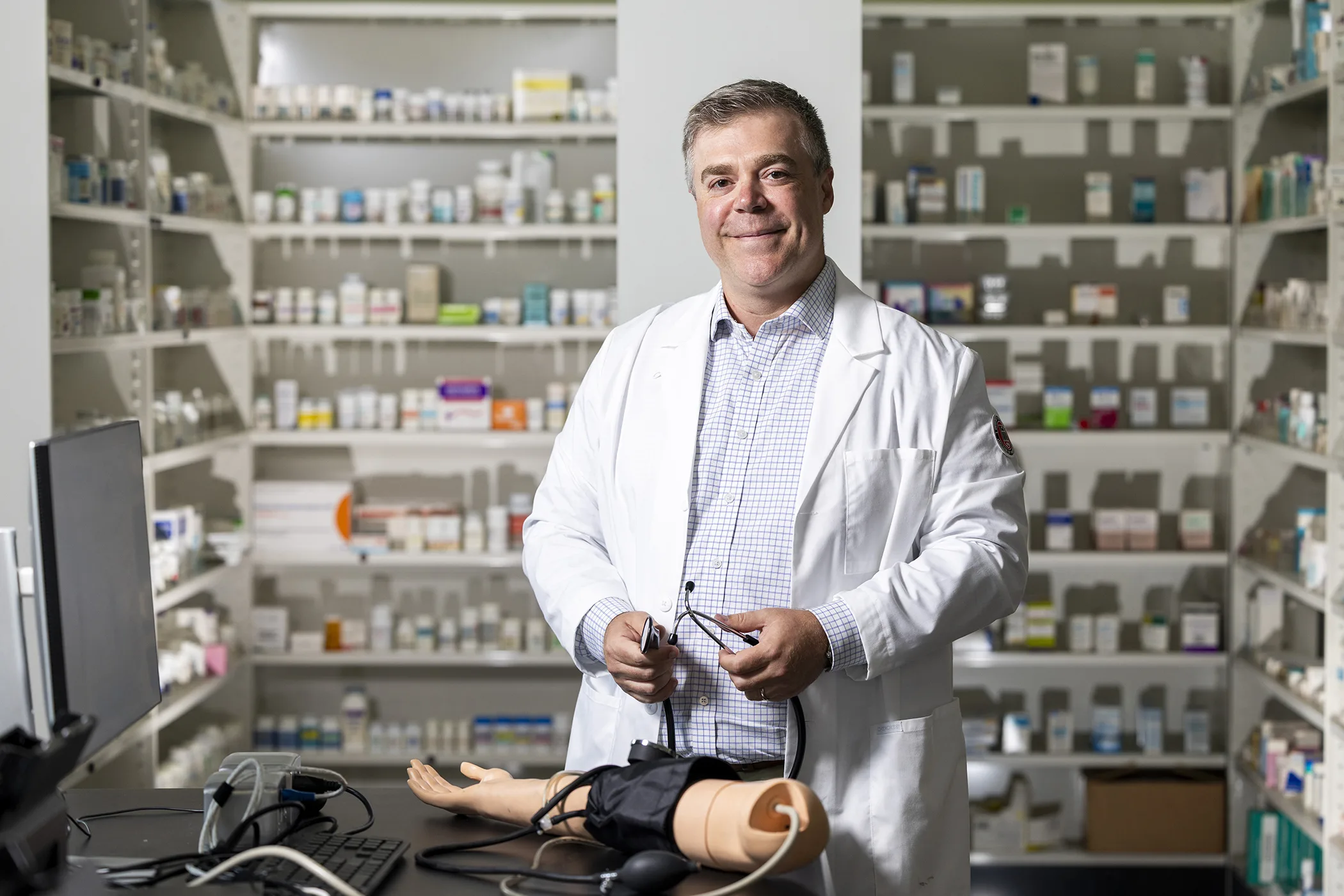 A pharmacist in a white coat stands in a simulation pharmacy with a stethoscope in his hands and blood pressure learning simulator in front of him