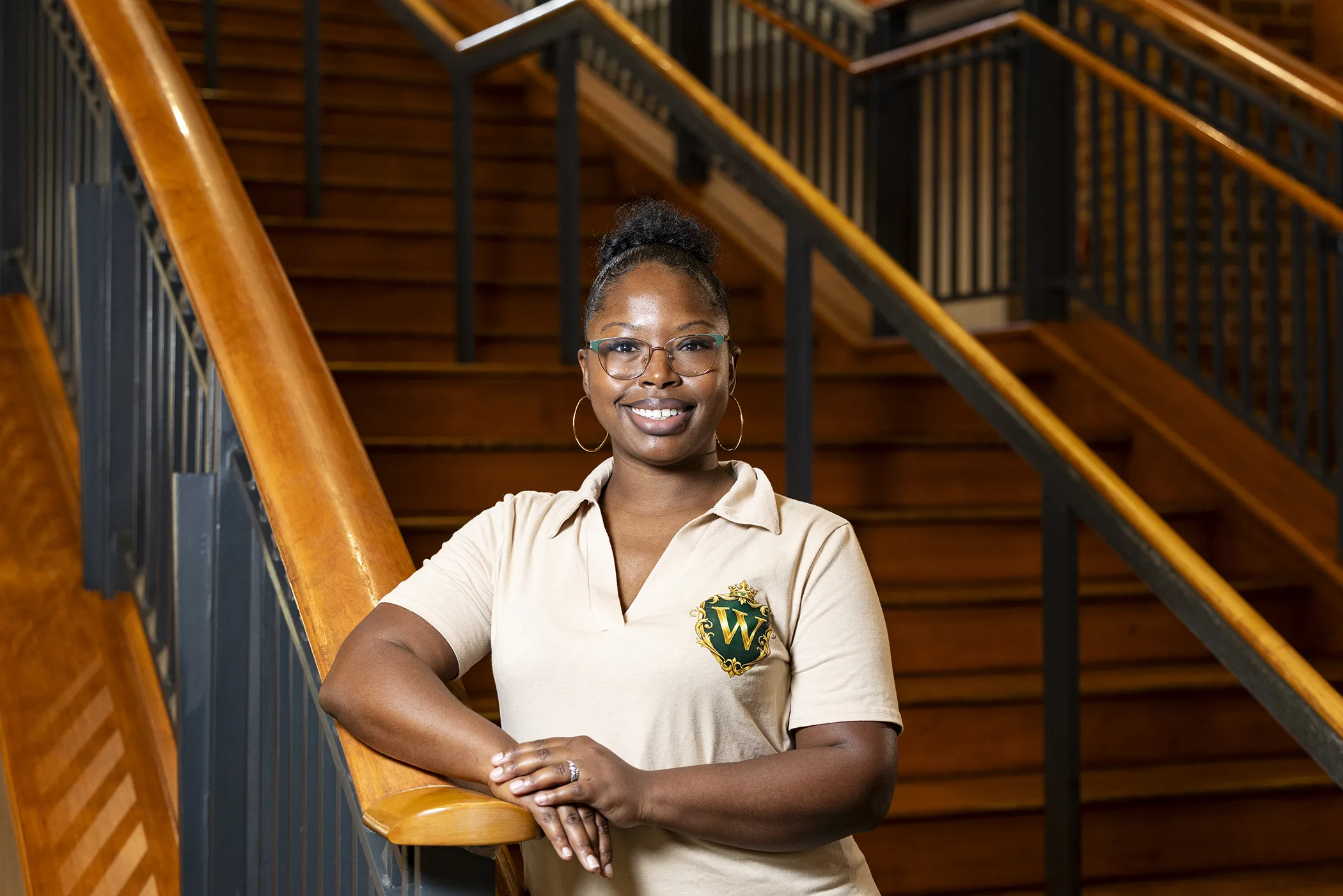 A woman stands at the bottom of a staircase with her right arm resting on the bannister