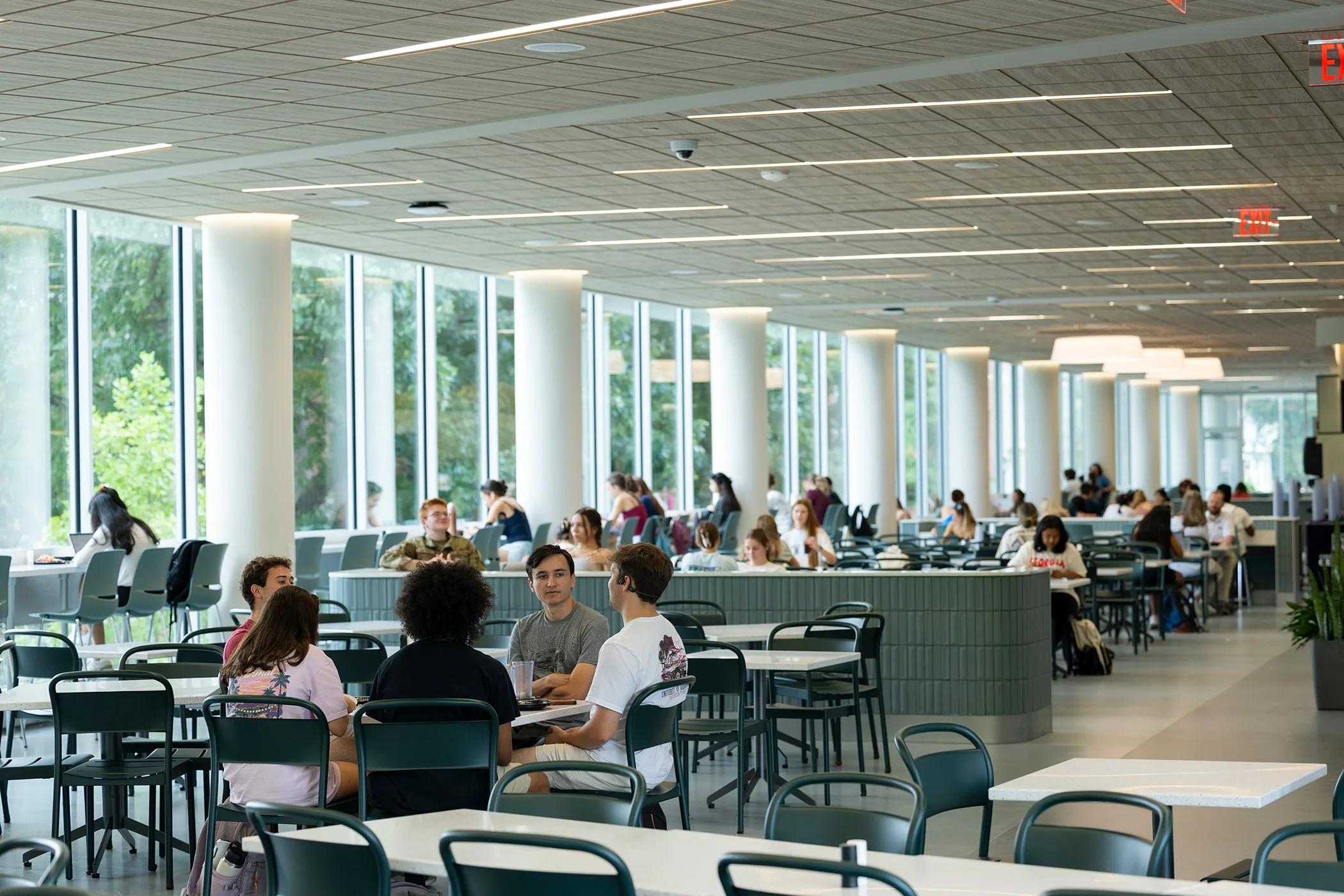 Students sit together in a dining hall as large windows fill the space with natural light and views of surrounding greenery.