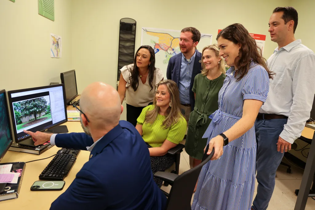 A group of people looks over a man’s shoulder as he points out information on a desktop computer.