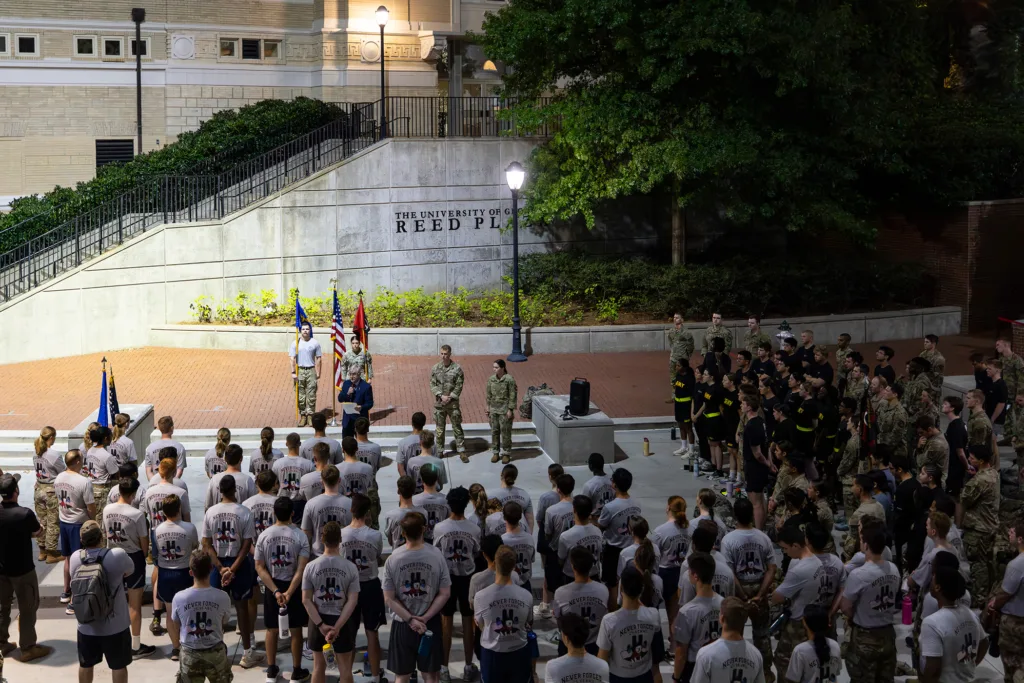 A crowd gathers before dawn at UGA’s Reed Plaza for a 9/11 remembrance ceremony, with ROTC cadets and firefighters standing at attention as a speaker addresses the group.