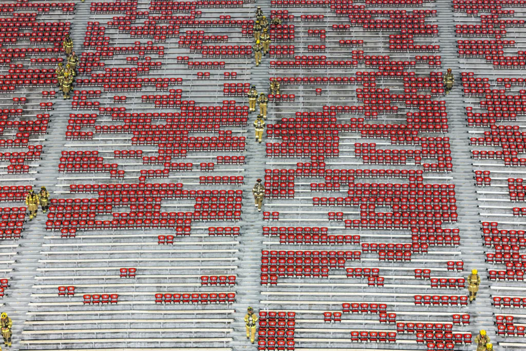 Multiple firefighters in full turnout gear climb stairs in a stadium filled with bleachers and red chairs.