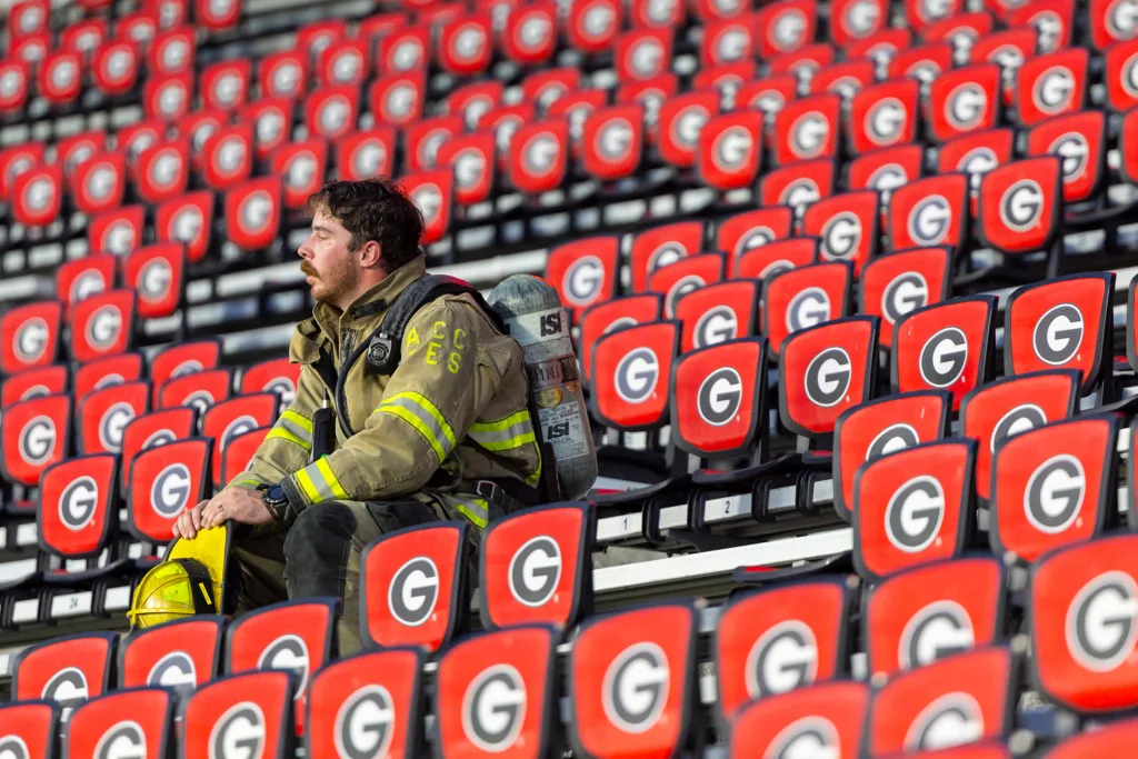 A firefighter in full turnout gear rests on a bleacher with red chairs behind him.