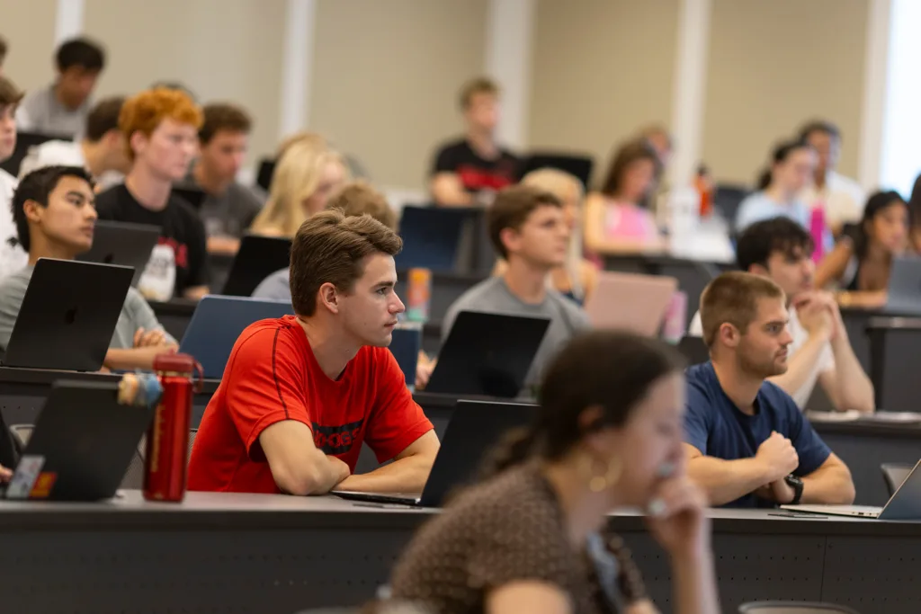 Students seated at classroom desks use laptops while listening to an instructor at the front of the room.