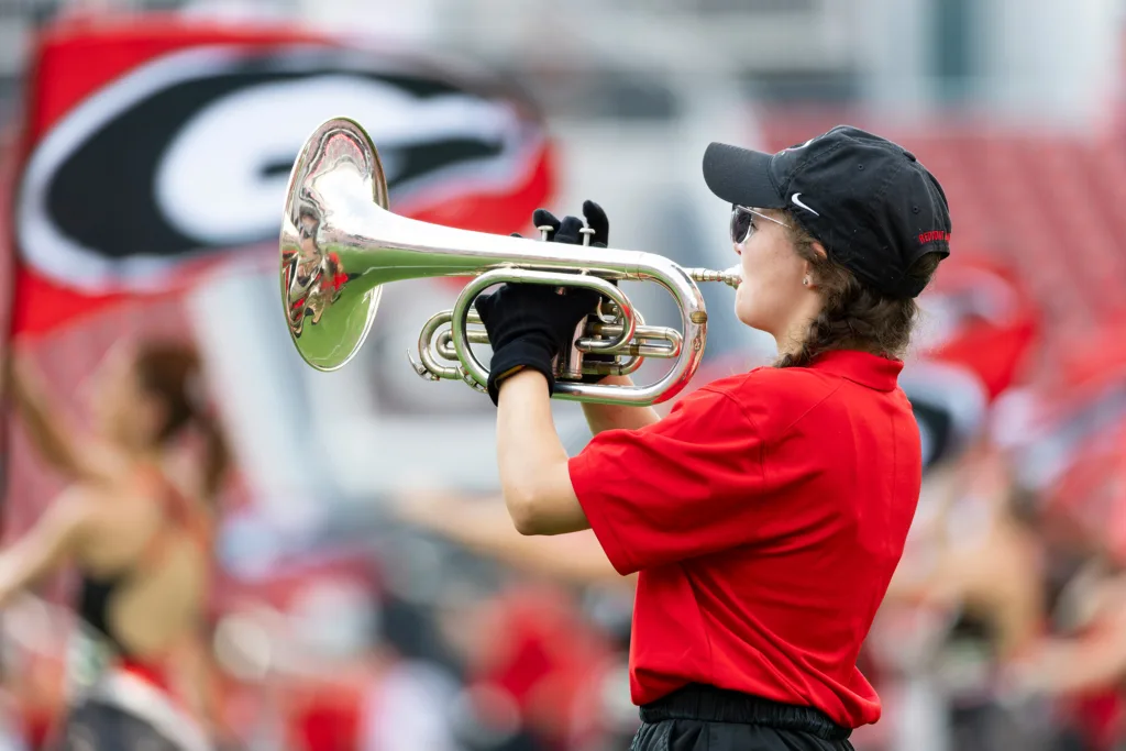 A young woman wearing a red polo shirt, a black cap and black gloves plays a golden trumpet. The background is a blurred crowd on a football field.