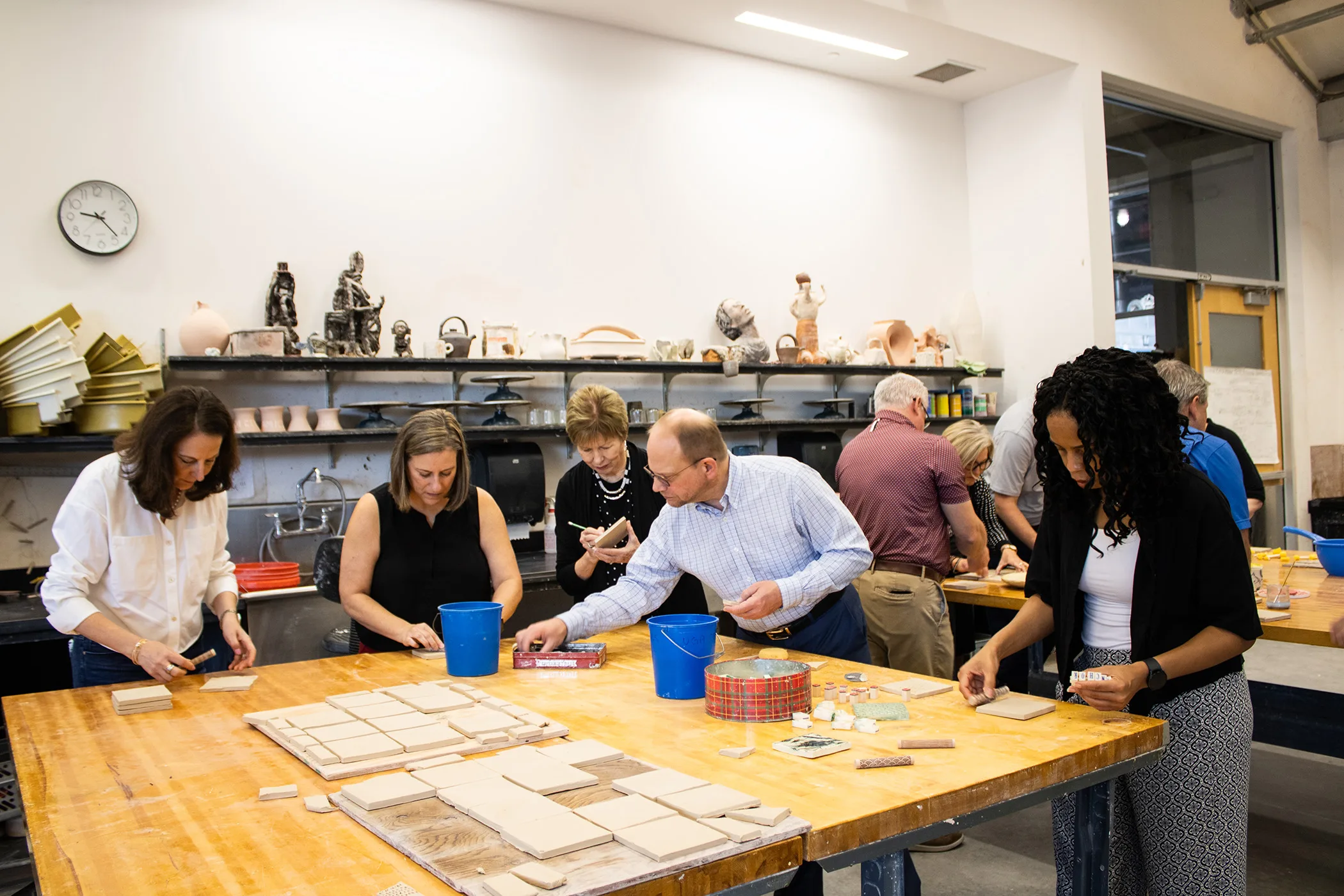 People gather around wooden tables and decorate clay tiles using pencils and other tools.