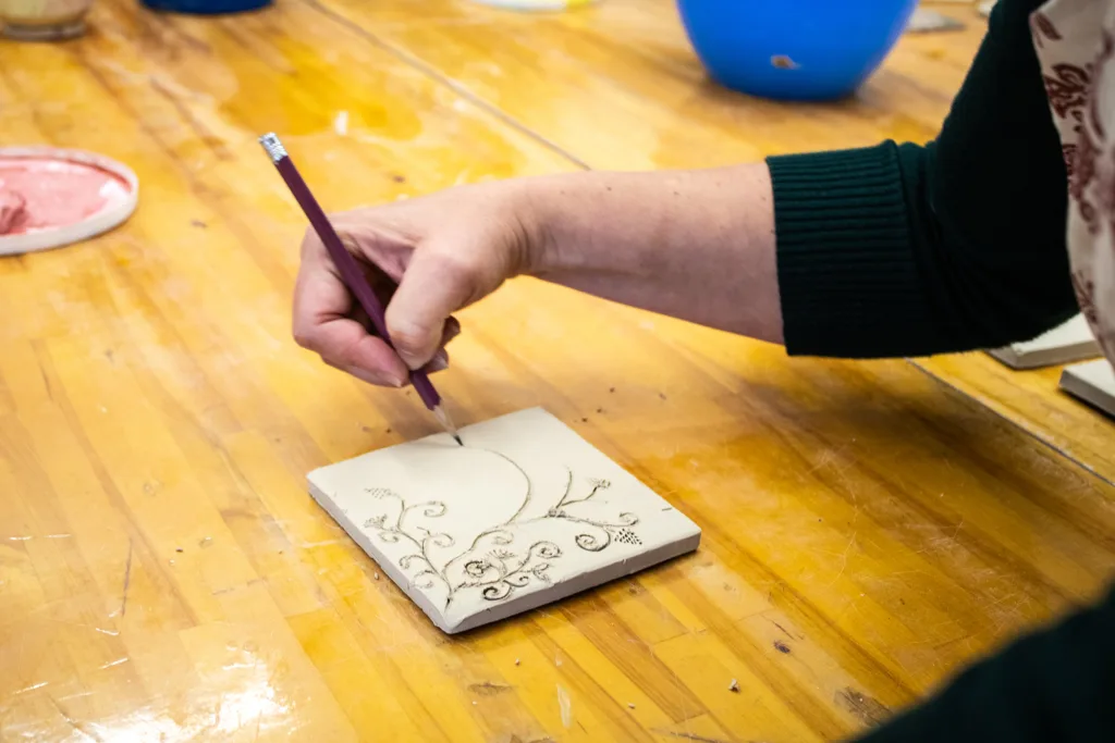 A person carves a decorative floral pattern on a clay tile using a pencil.