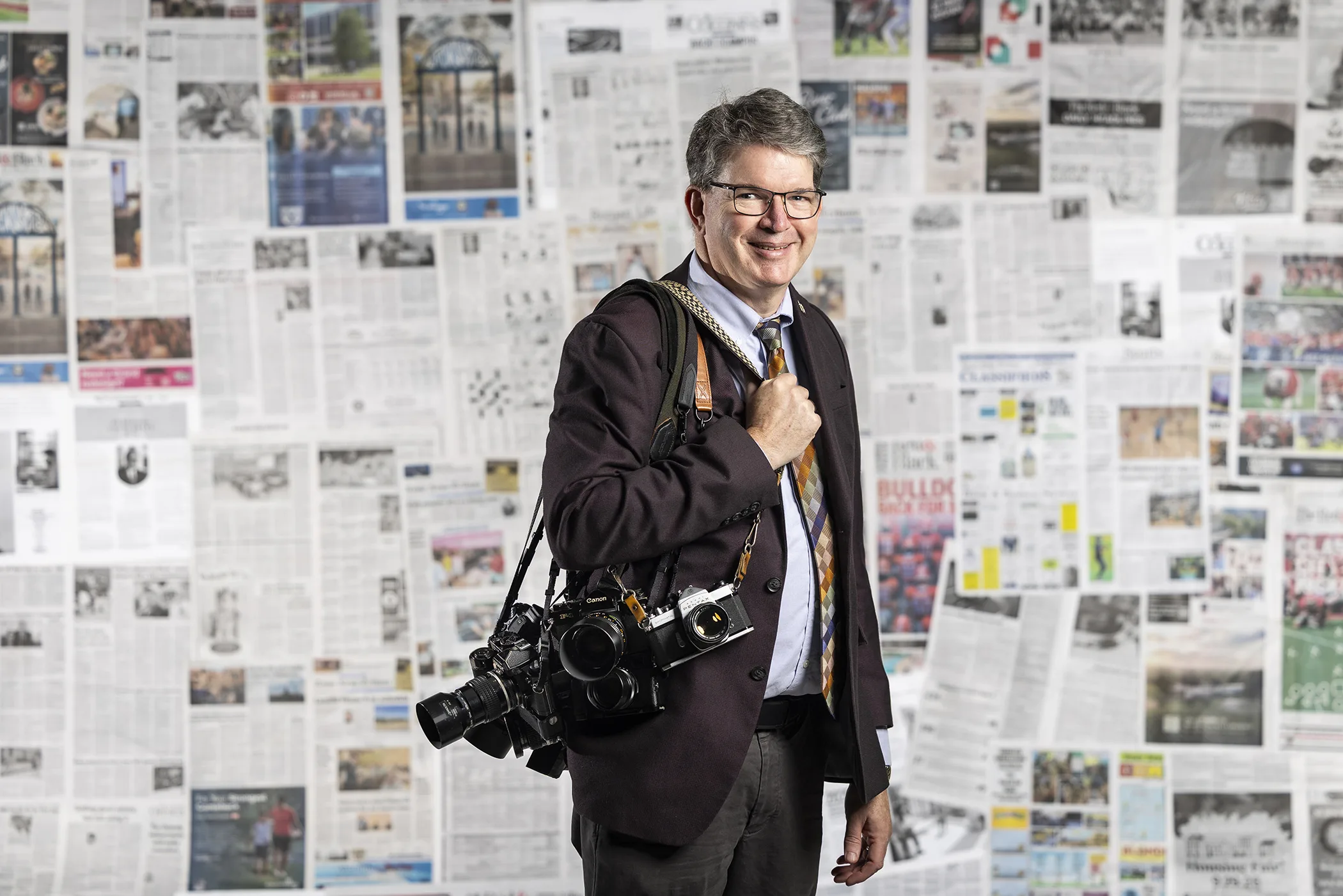 Man stands with numerous cameras over his shoulder in a room filled with newspapers.