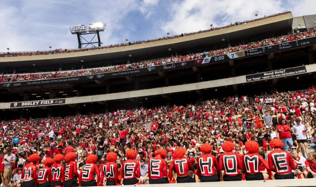 Students in red body paint and red wigs stand in a row with letters on their backs spelling "Home Sweet Home" in front of a packed student section at Sanford Stadium.