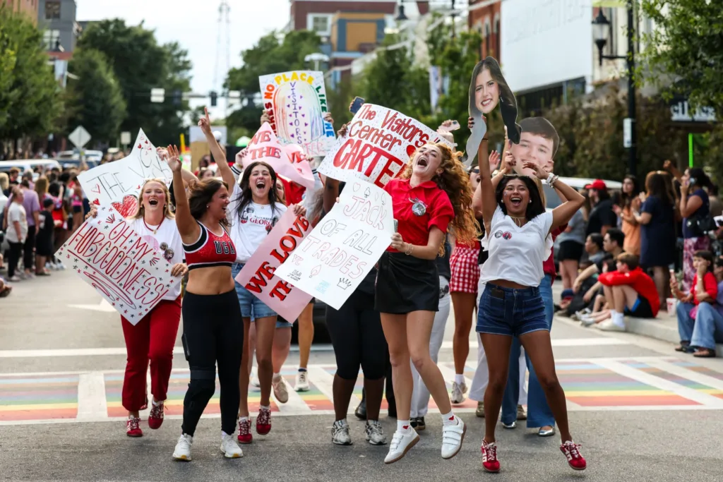 Students jump and cheer in a downtown Athens street during the homecoming parade, holding handmade signs and large cutout photos supporting homecoming court candidates.